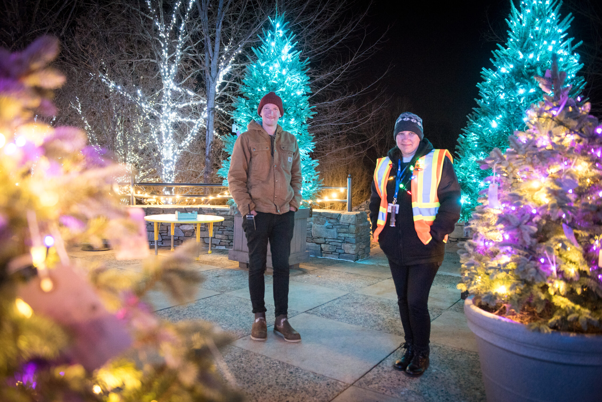 A Winter Lights staff member and volunteer stand in front of decorated holiday trees